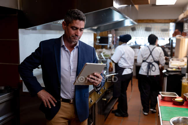 Front view close up of a middle aged Caucasian male restaurant manager standing with his hand on his hip using a tablet computer in a busy restaurant kitchen, while kitchen staff work in the background — Stock Photo