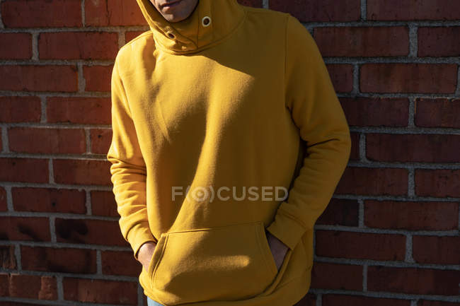 Vorderseite Mittelteil eines jungen Mannes mit gelbem Kapuzenpullover, der sich mit den Händen in der Vordertasche an eine Ziegelwand lehnt — Stockfoto