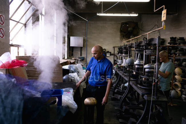 Side view of a middle aged mixed race man and a senior man operating equipment in the workshop at a hat factory, with steam and materials in the foreground — Stock Photo