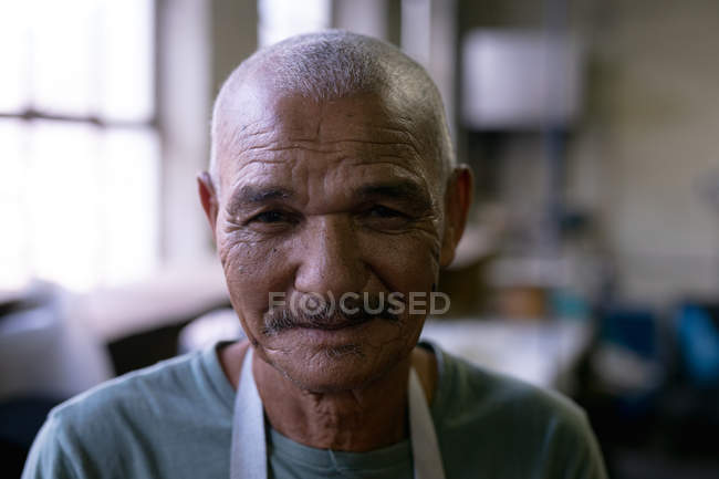 Retrato de cerca de un hombre de raza mixta senior mirando directamente a la cámara y sonriendo en el taller en una fábrica de sombreros - foto de stock