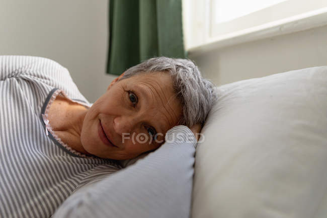 Вид спереди крупным планом зрелой кавказской женщины с короткими седыми волосами, лежащей на боку в постели дома, смотрящей в камеру и улыбающейся — стоковое фото