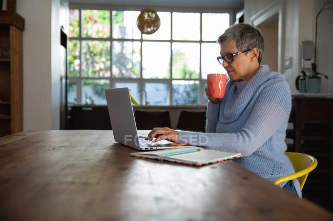 Seitenansicht einer reifen kaukasischen Frau mit kurzen grauen Haaren, die eine Brille trägt und an ihrem Esstisch sitzt, eine Tasse Kaffee hält und einen Laptop benutzt — Stockfoto