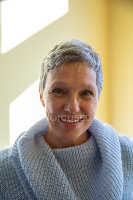 Retrato de cerca de una mujer caucásica madura con el pelo gris corto usando un suéter cuello capucha, mirando directamente a la cámara y sonriendo, con la luz del sol en la pared en el fondo - foto de stock