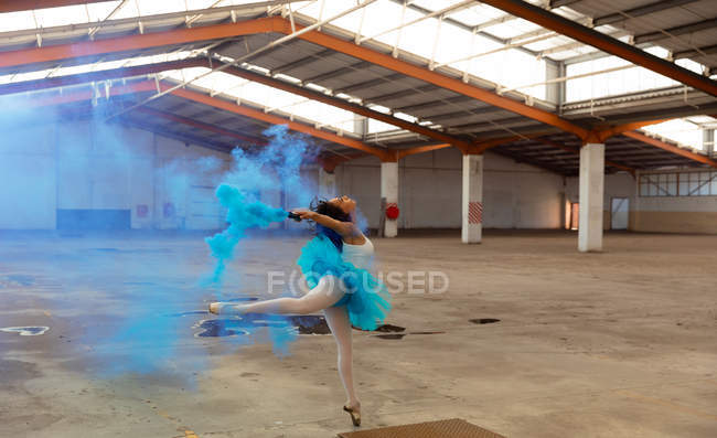 Vista lateral de uma jovem mista dançarina de balé usando um tutu azul e sapatos pontiagudos dançando segurando uma granada de fumaça azul em um quarto vazio em um armazém abandonado — Fotografia de Stock