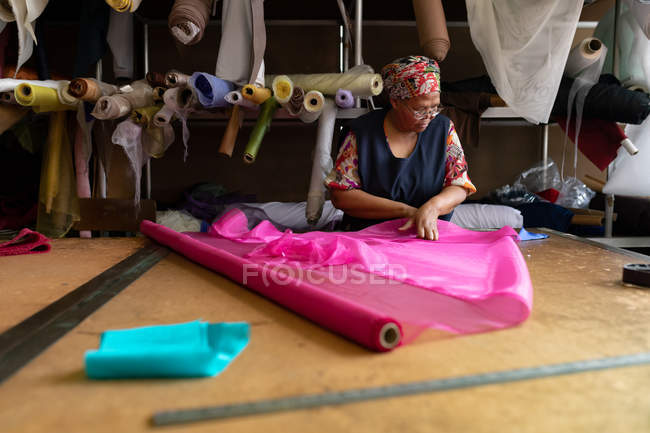 Vista frontale di una donna di razza mista di mezza età in piedi a un tavolo che lavora con tessuto rosa in una fabbrica di cappelli . — Foto stock