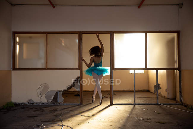Вид спереду молодої змішаної раси жіночої балетної танцівниці в блакитному взутті, що танцює у дверному отворі в покинутій будівлі складів, підсвічування сонячним світлом — стокове фото
