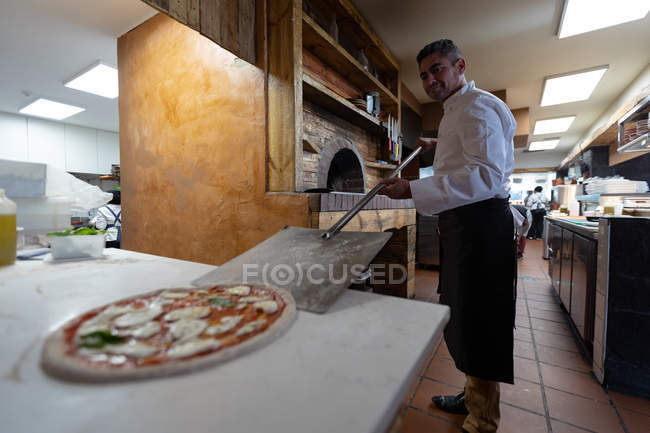 Вид сбоку крупным планом белого шеф-повара средних лет, берущего пиццу с кожурой от пиццы, чтобы поместить ее в духовку на кухне ресторана — стоковое фото