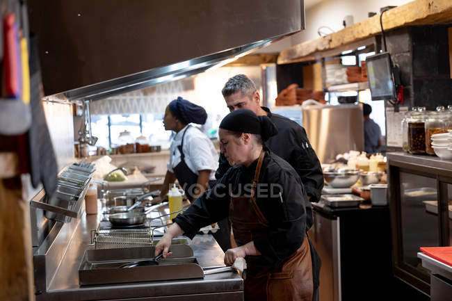 Seitenansicht von einem kaukasischen Koch mittleren Alters, der die Arbeit einer jungen kaukasischen Köchin überwacht, an der Bratstation in einer belebten Restaurantküche, während andere Küchenmitarbeiter im Hintergrund arbeiten — Stockfoto