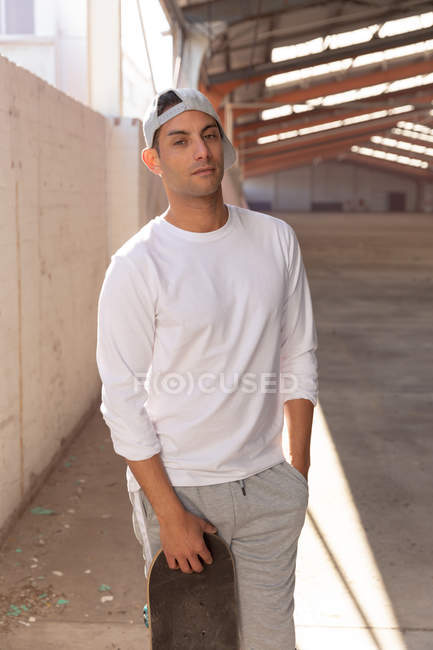 Front view close up of a young man wearing a baseball cap and holding a skateboard standing with one hand in his pocket, looking to camera, in an abandoned warehouse — Stock Photo