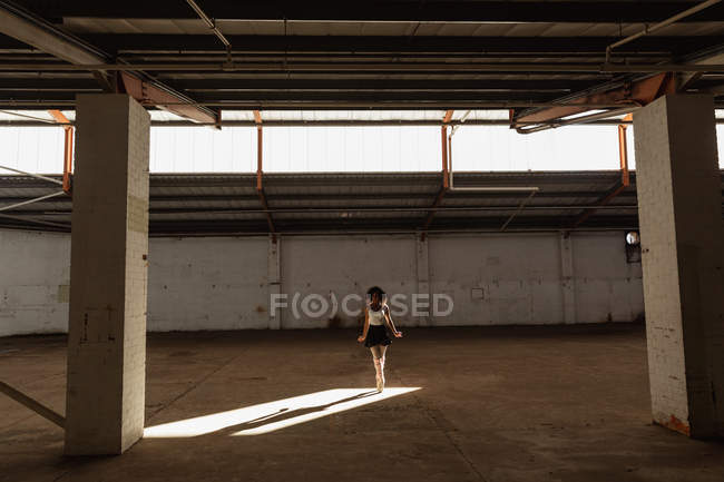 Veduta frontale di una giovane ballerina di danza mista che indossa scarpe a punta in piedi sulle dita dei piedi in un pozzo di luce solare con le braccia ai lati mentre balla in una stanza vuota in un magazzino abbandonato — Foto stock