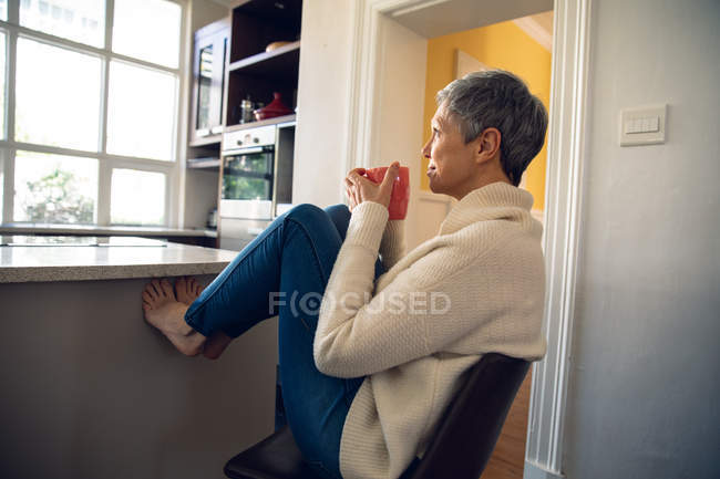 Вид сбоку на взрослую белую женщину с короткими седыми волосами, сидящую на стуле на кухне с поднятыми ногами, пьющую чашку кофе и выглядывающую в окно — стоковое фото