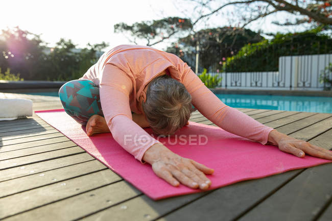 Front view low section of a mature Caucasian woman wearing sports clothes sitting on a mat leaning forward in a yoga position, exercising by the swimming pool in her garden, back lit by sunlight with a rural view in the background — Stock Photo