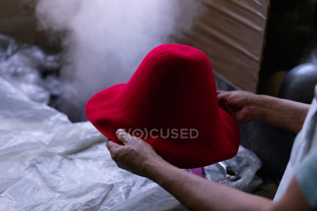 Sobre la vista del hombro de las manos del hombre sosteniendo la parte superior de un sombrero rojo que se ha formado en una pieza de equipo en el taller en una fábrica de sombreros, con vapor y materiales en el fondo - foto de stock