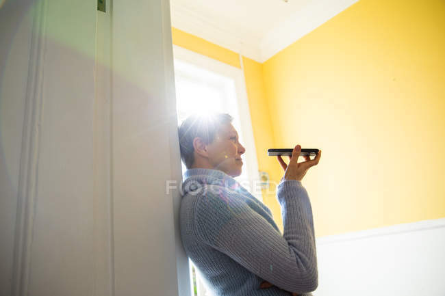 Side view of a mature Caucasian woman with short grey hair leaning on a wall at home using a smartphone she is holding in front of her and smiling, back lit by sunlight — Stock Photo