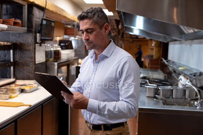 Side view close up of a middle aged Caucasian male restaurant manager writing on a clipboard in a restaurant kitchen — Stock Photo