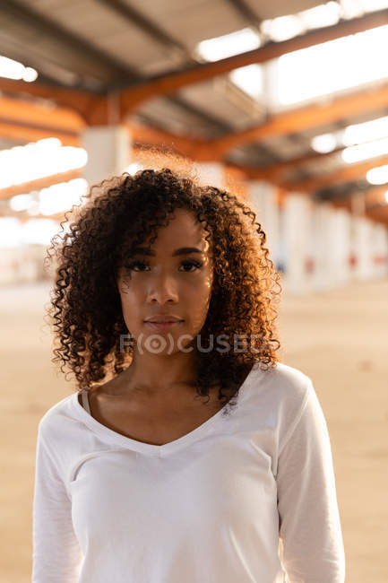 Портрет крупним планом молодої змішаної раси жінки з довжиною плеча кучеряве волосся дивиться прямо на камеру в покинутому складі — стокове фото