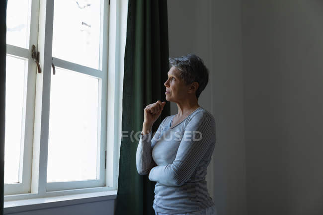 Vista lateral de uma mulher branca madura com cabelos brancos curtos em pé e olhando para fora da janela em casa — Fotografia de Stock