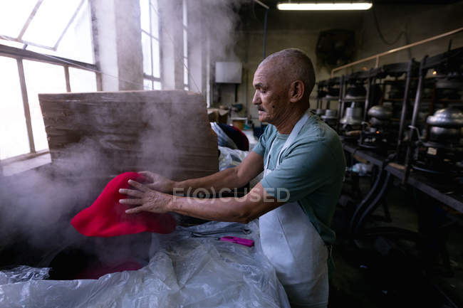 Вид сбоку пожилого мужчины смешанной расы в фартуке, который держит в руках верхушку шляпы, прожаренной в мастерской на шляпной фабрике, на заднем плане видно оборудование — стоковое фото