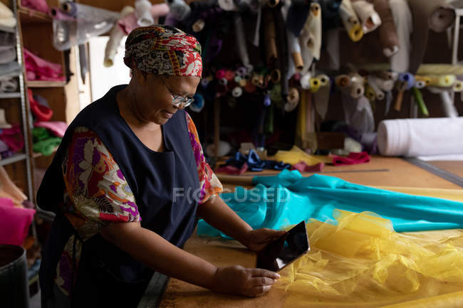 Vista laterale da vicino di una donna di razza mista di mezza età in piedi a un tavolo con tessuto blu e giallo su di esso utilizzando un computer tablet mentre si lavora in una fabbrica di cappelli . — Foto stock