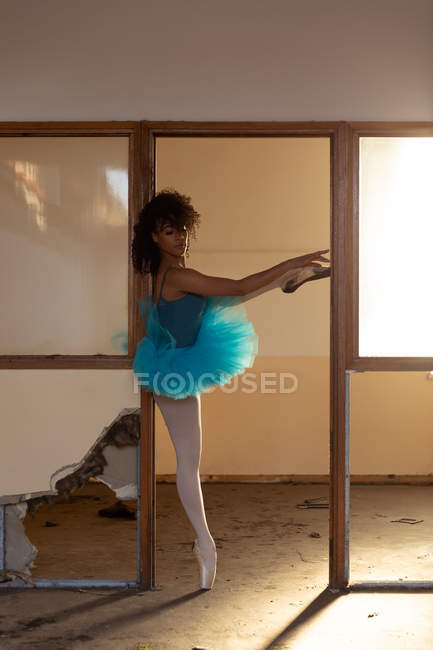Vue latérale d'une jeune danseuse de ballet mixte portant un tutu bleu et des chaussures de pointe debout sur une jambe sur ses orteils dans une porte d'un entrepôt abandonné — Photo de stock