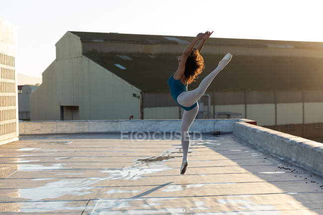 Вид збоку на молоду змішану гонку танцівниця балету, що стрибає руками і піднята ногою, на даху міської будівлі, підсвічування сонячним світлом — стокове фото