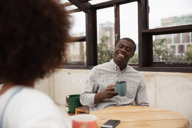 За плечом молодой женщины смешанной расы и молодого афроамериканца, сидящего за столом, пьющего кофе и разговаривающего в стеклянной крытой комнате на крыше, с городскими зданиями на заднем плане — стоковое фото