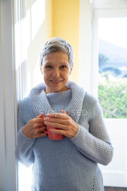 Retrato de cerca de una mujer caucásica madura con el pelo gris corto usando un suéter cuello capucha, de pie delante de una ventana en casa sosteniendo una taza de café, mirando directamente a la cámara y sonriendo, la luz del sol en el fondo - foto de stock