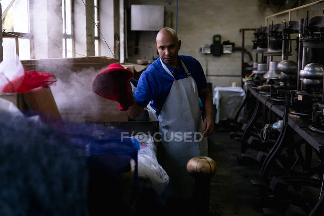 Front view of a middle aged mixed race man wearing an apron operating a piece of equipment in the workshop at a hat factory, with steam and materials in the foreground and equipment in the background — Stock Photo