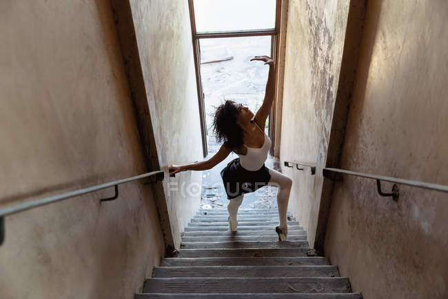Vista lateral elevada de uma jovem dançarina de balé mista dançando em uma escadaria em um armazém abandonado — Fotografia de Stock