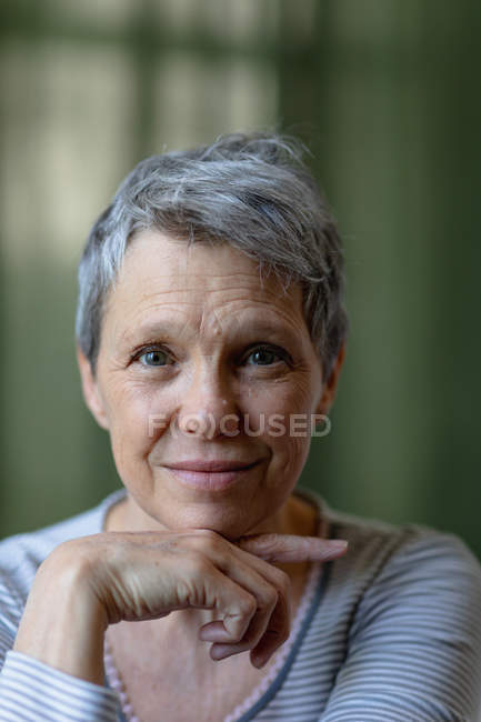 Portrait close up of a mature Caucasian woman with short grey hair looking straight to camera and smiling, with her chin resting on her hand — Stock Photo