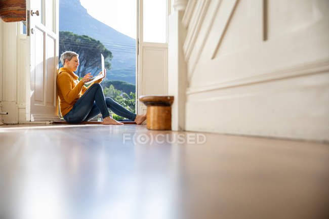 Surface level side view of a mature Caucasian woman sitting on the floor in the doorway of her house in the sun using a laptop computer, with sunlit trees outside in the background, seen from the hallway of her house — Stock Photo