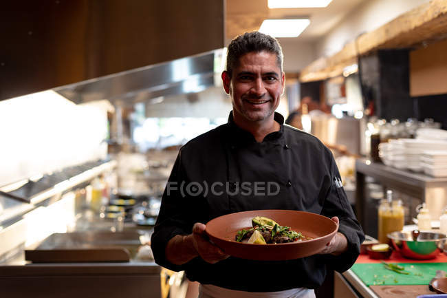 Portrait d'un homme caucasien souriant d'âge moyen tenant un plat de nourriture préparée dans une cuisine de restaurant — Photo de stock