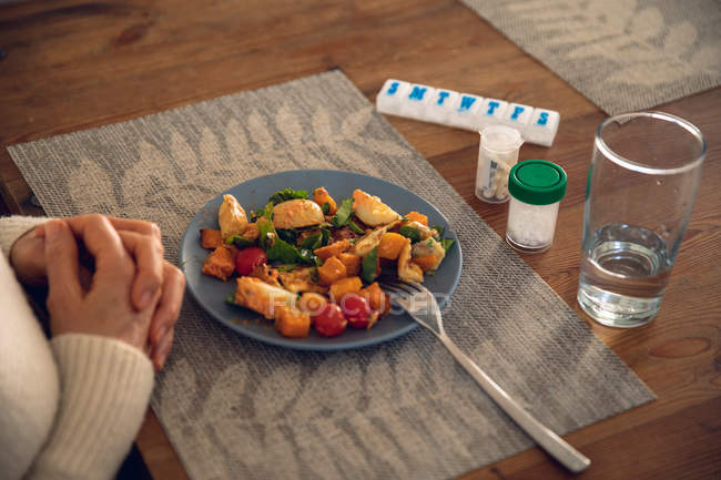 Primer plano de las manos de la mujer que descansa sobre la mesa, con comida en un plato, frascos de tabletas y pastillas y un vaso de agua frente a ella - foto de stock