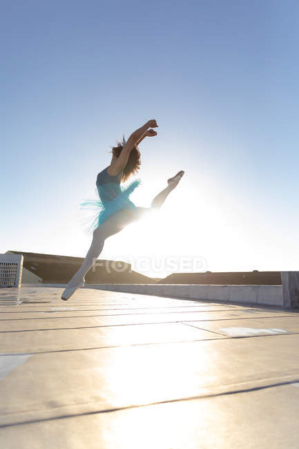 Seitenansicht einer jungen Balletttänzerin mit gemischter Rasse, die ein blaues Tutu und Spitzenschuhe trägt und mit erhobenem Arm auf dem Dach eines städtischen Gebäudes im Gegenlicht der Sonne springt — Stockfoto