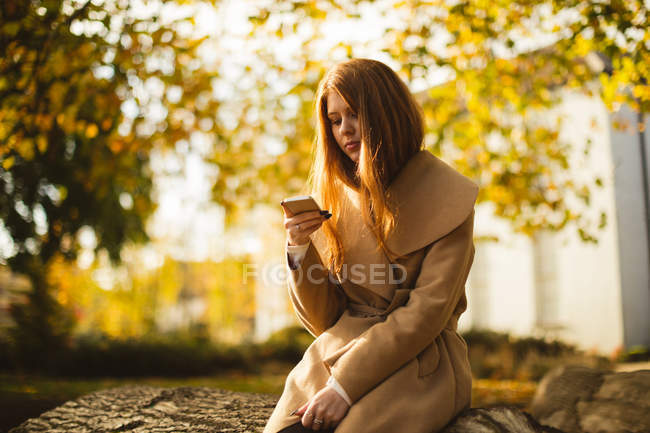 Mujer usando el teléfono móvil mientras está sentado en un tronco de árbol en el parque . - foto de stock