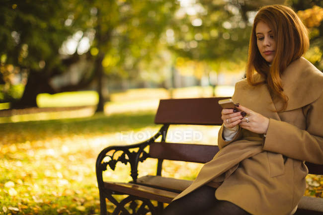 Жінка використовує мобільний телефон, сидячи на лавці в парку . — стокове фото