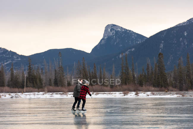 Vista lateral de la joven pareja caucásica patinando juntos en un paisaje nevado natural - foto de stock
