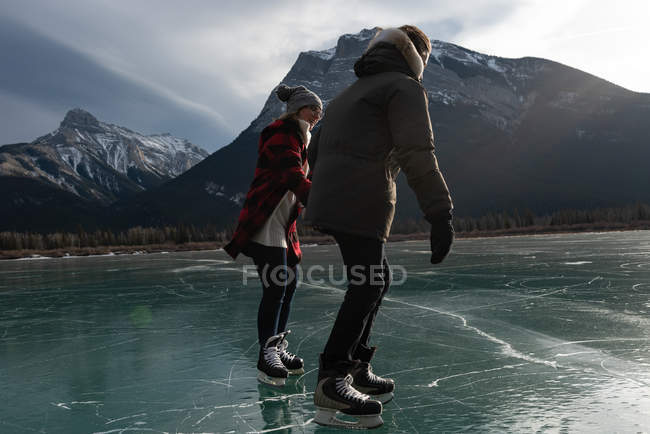 Vista lateral de la joven pareja caucásica patinando juntos en el paisaje nevado natural en el fondo - foto de stock