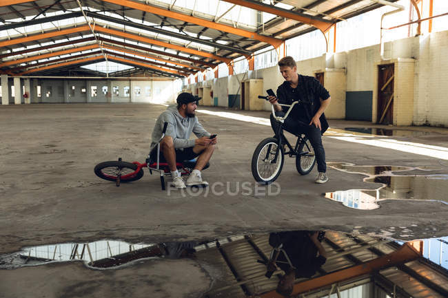 Frontansicht von zwei jungen erwachsenen kaukasischen Männern, die auf BMX-Fahrrädern sitzen, miteinander reden und Smartphones in einer verlassenen Lagerhalle benutzen — Stockfoto