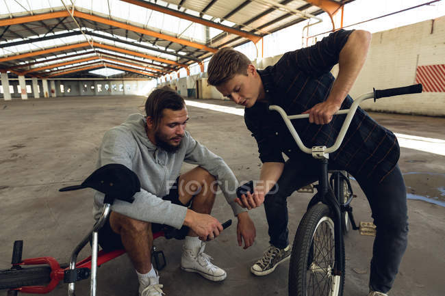 Nahaufnahme von zwei jungen kaukasischen Männern, die auf BMX-Fahrrädern sitzen und reden, wobei der eine dem anderen sein Smartphone in einer verlassenen Lagerhalle zeigt. — Stockfoto