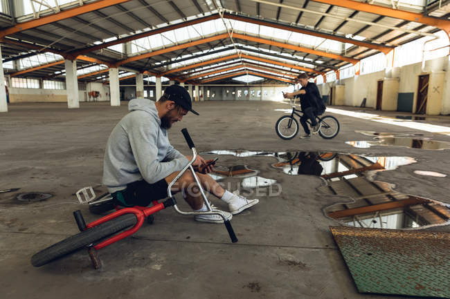 Вид сбоку на двух молодых взрослых белых мужчин, сидящих на велосипедах BMX с помощью смартфонов на заброшенном складе — стоковое фото