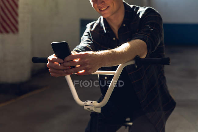 Vorderansicht Mittelteil eines lächelnden jungen kaukasischen Mannes, der auf einem BMX-Fahrrad sitzt, sich an den Lenker lehnt und ein Smartphone in einer verlassenen Lagerhalle benutzt — Stockfoto