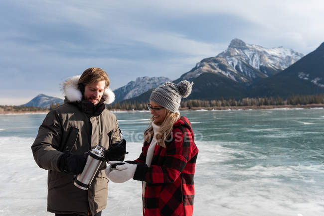 Vista frontal do casal caucasiano feliz tomando café enquanto está em pé na paisagem nevada natural no fundo — Fotografia de Stock