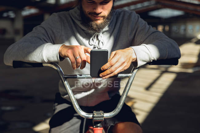 Vista frontale da vicino di un giovane caucasico con la barba seduto su una bici BMX, appoggiato al manubrio e con uno smartphone in un magazzino abbandonato — Foto stock