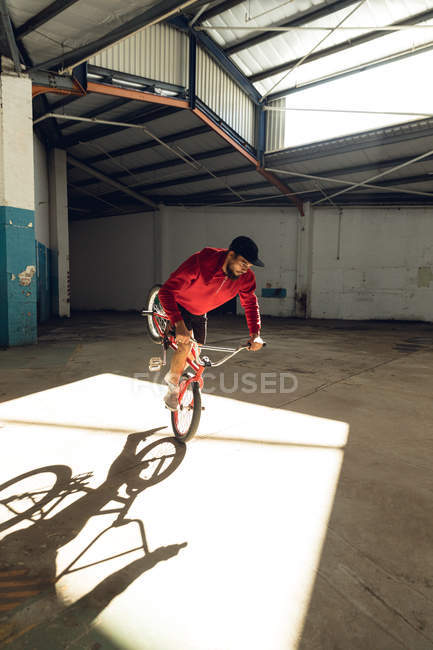Vista frontal de um jovem caucasiano balanceando na roda dianteira de uma bicicleta BMX em um eixo de luz solar enquanto pratica truques em um armazém abandonado — Fotografia de Stock