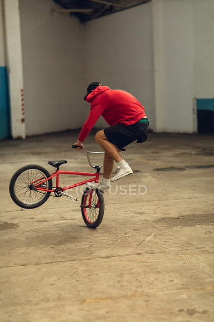 Vista lateral de perto de um jovem caucasiano vestindo shorts, um top com capuz e treinadores realizando um truque de pé na roda dianteira de uma bicicleta BMX em um armazém abandonado — Fotografia de Stock