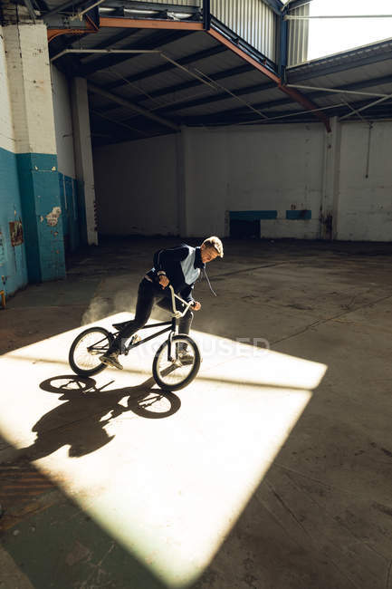 Side view close up of a young Caucasian man sliding sideways to halt in a shaft of sunlight on a BMX bike while practicing tricks in an abandoned warehouse — Stock Photo