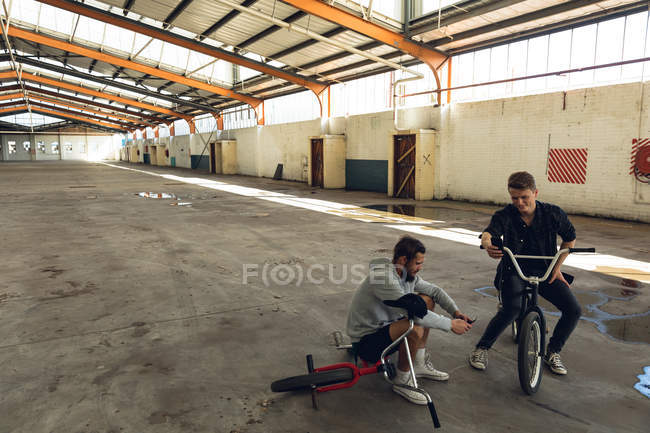 Вид спереди на двух молодых взрослых белых мужчин, сидящих на велосипедах BMX, разговаривающих друг с другом и использующих смартфоны на заброшенном складе — стоковое фото