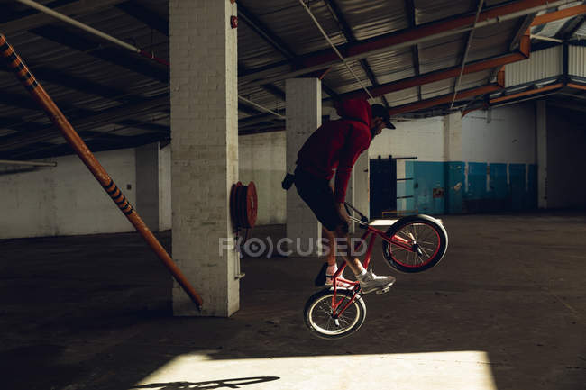 Vista lateral de um jovem caucasiano de pé e pulando em uma bicicleta BMX em um eixo de luz solar enquanto pratica truques em um armazém abandonado — Fotografia de Stock