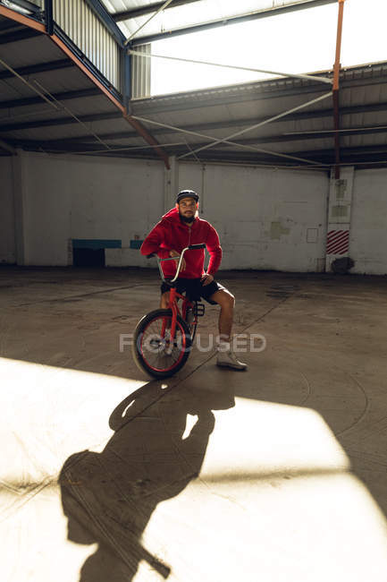Porträt eines jungen kaukasischen Mannes mit Bart, Baseballmütze, kurzer Hose und rotem Oberteil, der in einer verlassenen Lagerhalle auf einem BMX-Fahrrad sitzt und in die Kamera blickt, während das Sonnenlicht seinen Schatten vor sich wirft. — Stockfoto